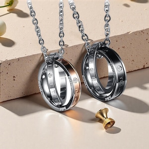 Pode incluir: Dois colares com anéis entrelaçados prateados. Um anel tem uma faixa em ouro rosa, o outro é preto. Cada anel tem as palavras "Forever" e "Eternity" gravadas. Um pequeno objeto decorativo dourado na parte inferior.