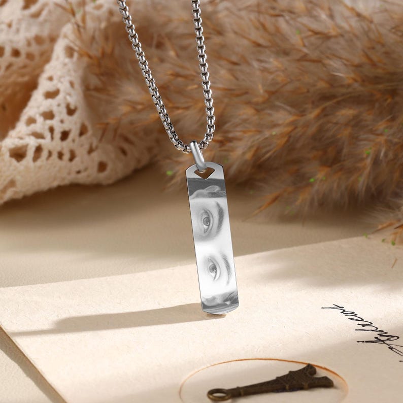 Puede incluir: Un collar plateado con un colgante rectangular que muestra una imagen en escala de grises de dos ojos. El colgante cuelga de una cadena fina. El fondo incluye tela beige y plantas secas.