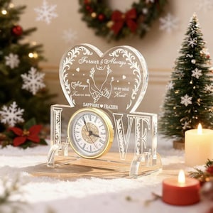 Könnte beinhalten: Eine transparente Acryluhr mit goldfarbenem Zifferblatt und dem Wort "LOVE" in der Basis. Die Uhr hat eine herzförmige Oberseite mit den Worten "Forever & Always" und anderem Text. Die Uhr ist ein dekorativer Artikel.