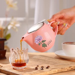 May include: A pink ceramic teapot with a silver lid is pouring a dark liquid into a clear glass cup. The teapot has a floral design and the name "Rachel" printed on it. Coffee beans are scattered on the wooden surface.