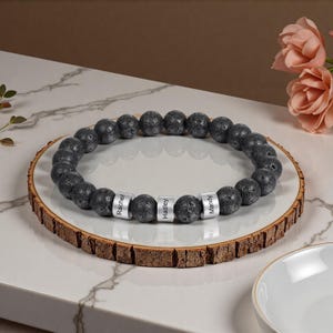 Könnte beinhalten: Ein schwarzes Lavastein-Armband mit drei silbernen Namensperlen mit den Aufschriften "Rachel", "Hadley" und "Mandy". Das Armband liegt auf einem weißen Teller mit Holzrand.