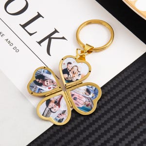 May include: A gold-colored, four-leaf clover keychain with heart-shaped photo frames. Each frame contains a color photograph. The keychain has a circular ring for attaching keys. The background includes the text "OLK" and "KE AND DO".