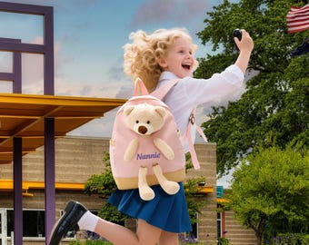 May include: A pink backpack with a teddy bear design and the name "Nannie" embroidered on the front. The backpack has a light brown trim and straps. The child is wearing a white shirt, blue skirt, and black shoes.