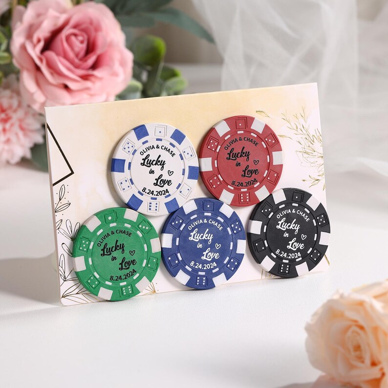 May include: A set of five poker chip favors with the text "Olivia & Chase Lucky in Love 8.24.2024". The chips are white, red, green, blue, and black with white text and a heart symbol. The chips are arranged on a decorative card.