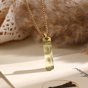 Puede incluir: Collar dorado con un colgante rectangular con un dise&ntilde;o de ojos. El colgante cuelga de una cadena fina. El fondo presenta tonos y texturas suaves y neutros.