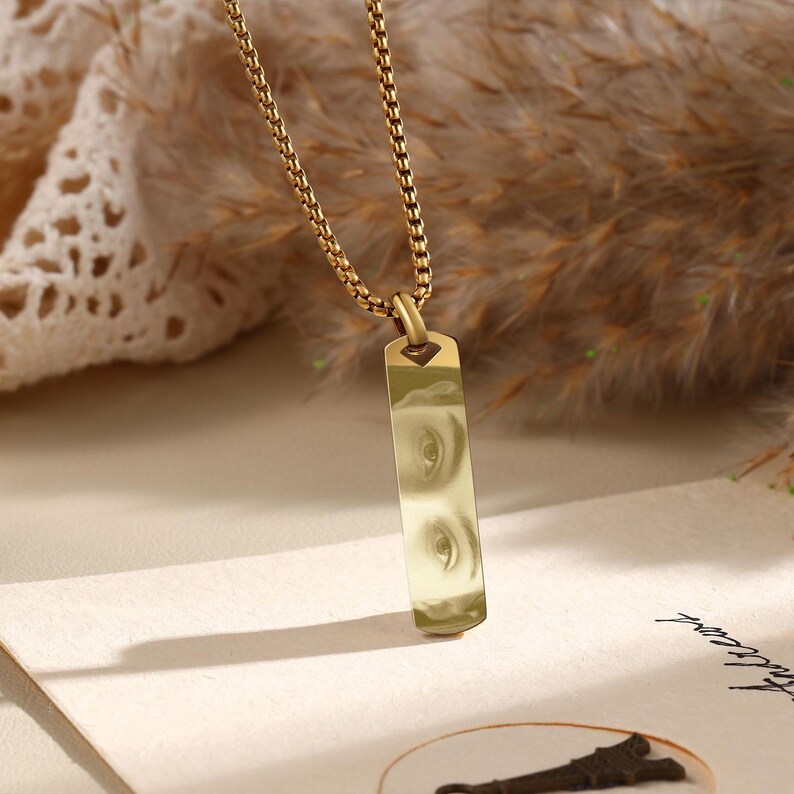 Puede incluir: Collar dorado con un colgante rectangular con un dise&ntilde;o de ojos. El colgante cuelga de una cadena fina. El fondo presenta tonos y texturas suaves y neutros.