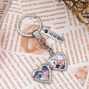 Op de afbeelding: Zilverkleurige sleutelhanger met twee hartvormige medaillons met foto's. Een rechthoekig label met een muzieknoot en een barcode-achtig ontwerp hangt boven de medaillons. De sleutelhanger ligt op een bedrukte papieren achtergrond.