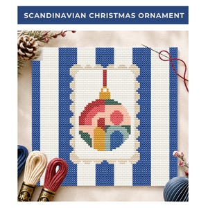 Op de afbeelding: Een kruissteekpatroon met een centraal kerstornamentontwerp met geometrische vormen in roze, rood, geel en groen, omlijst door een geschulpte rand op een blauw-witte gestreepte achtergrond, met de tekst SCANDINAVIAN CHRISTMAS ORNAMENT.