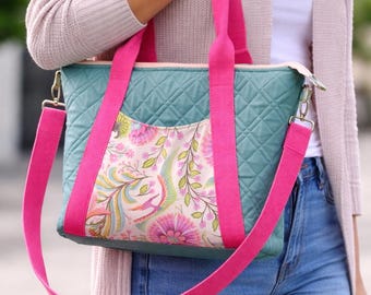 May include: A teal quilted tote bag with a floral print pocket and bright pink straps. The bag has a zippered top and a detachable shoulder strap. The bag is being carried by a person wearing a light pink cardigan and blue jeans.