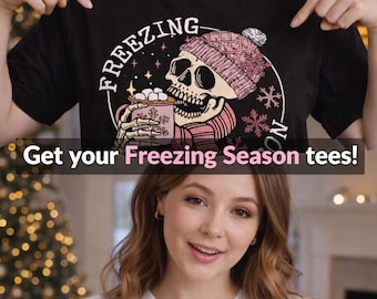 May include: Black and white t-shirt with a skull graphic wearing a pink beanie and scarf, holding a mug of marshmallows. The word "FREEZING" is arched above the skull. The text "Get your Freezing Season tees!" is below the graphic.