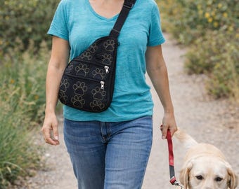 May include: Black sling bag with paw print design. The bag has three zippered compartments and a black strap. A person is wearing the bag while walking a yellow Labrador Retriever on a path.