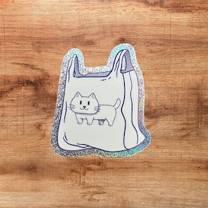 Könnte beinhalten: Ein holografischer Aufkleber in Form einer Plastiktüte mit einer dunkelblauen Strichzeichnung einer Katze auf der Vorderseite. Der Aufkleber hat eine schimmernde, mehrfarbige reflektierende Oberfläche, die das Licht einfängt und die Form betont.