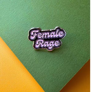 May include: An enamel pin with the words "Female Rage" in a cursive font, edged in silver. The pin is set against a green and yellow background.