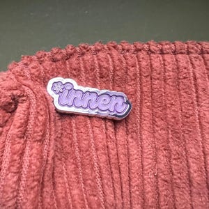 Op de afbeelding: Een zilverkleurige pin met het woord "innen" in paarse letters, geaccentueerd met een kleine bloem. De pin is geplaatst op een textiel met een corduroy-achtige structuur in een gedempte roodbruine kleur.