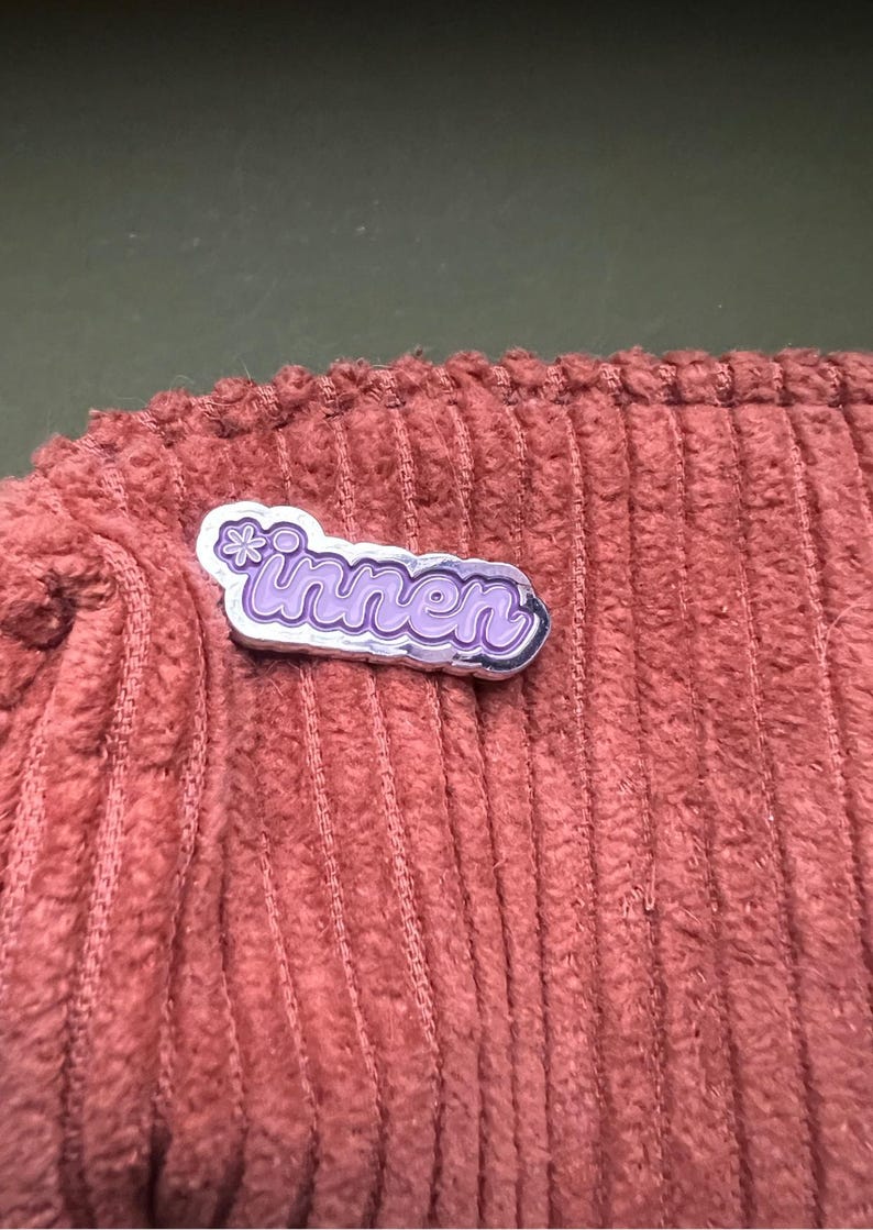 Op de afbeelding: Een zilverkleurige pin met het woord "innen" in paarse letters, geaccentueerd met een kleine bloem. De pin is geplaatst op een textiel met een corduroy-achtige structuur in een gedempte roodbruine kleur.