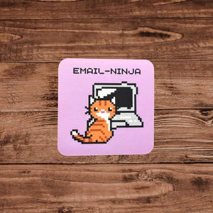 Könnte beinhalten: Ein quadratischer Untersetzer mit einer pixeligen orangefarbenen Tabby-Katze vor einem pixeligen Laptop. Der Text "EMAIL-NINJA" befindet sich über der Katze und dem Laptop auf einem rosa Hintergrund. Der Untersetzer liegt auf einer Holzoberfläche.