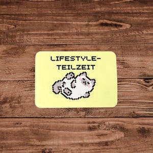Könnte beinhalten: Ein gelber rechteckiger Untersetzer mit dem Text "LIFESTYLE-TEILZEIT" und einer pixeligen Illustration einer Katze. Der Untersetzer liegt auf einem Holzgrund.