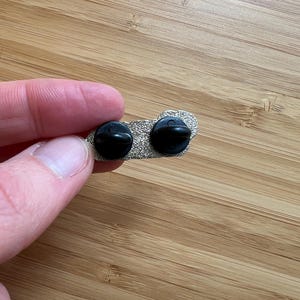 Op de afbeelding: Close-up van de achterkant van een zilverkleurige emaille pin met twee zwarte pin backs. De pin ligt op een houten oppervlak.