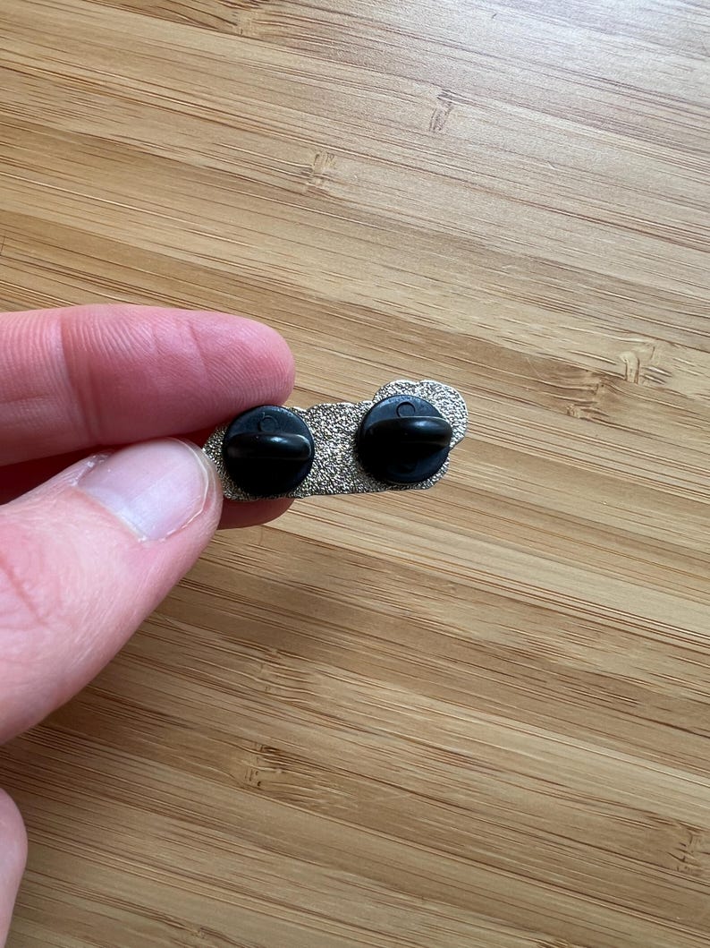 Op de afbeelding: Close-up van de achterkant van een zilverkleurige emaille pin met twee zwarte pin backs. De pin ligt op een houten oppervlak.