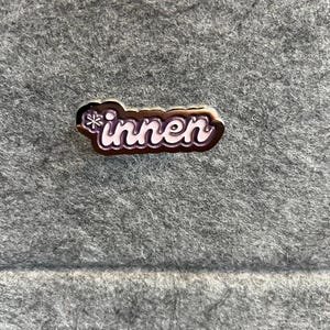 Op de afbeelding: Een rechthoekige emaille pin met het Duitse woord "innen" in witte scriptletters. De pin heeft een donkerbruine rand en een sneeuwvlokontwerp. De pin is geplaatst tegen een grijze vilt achtergrond.