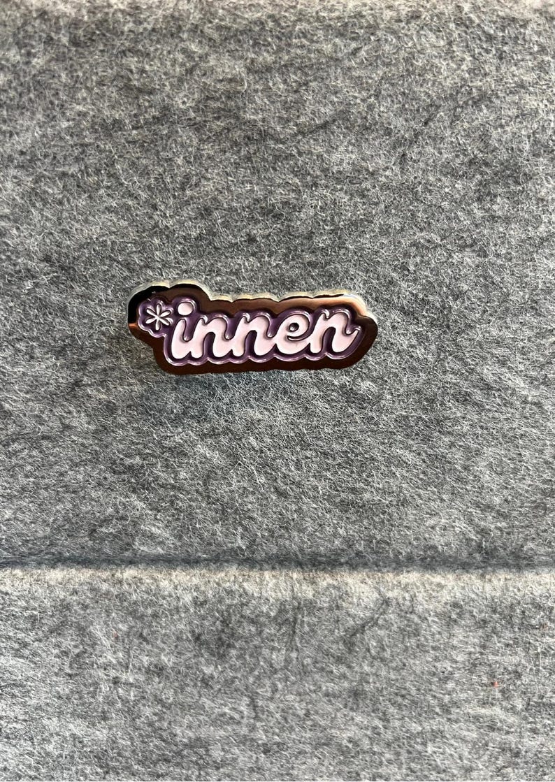 Op de afbeelding: Een rechthoekige emaille pin met het Duitse woord "innen" in witte scriptletters. De pin heeft een donkerbruine rand en een sneeuwvlokontwerp. De pin is geplaatst tegen een grijze vilt achtergrond.