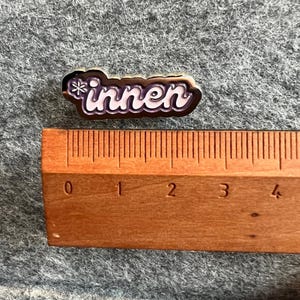 Op de afbeelding: Een kleine, rechthoekige speld met het Duitse woord "innen" in witte scriptletters, omrand in bruin en paars. Een houten liniaal ligt naast de speld en toont de afmetingen in centimeters.