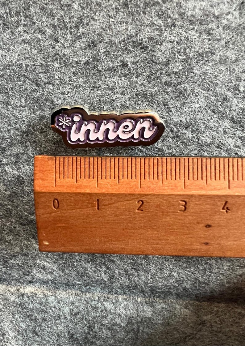 Op de afbeelding: Een kleine, rechthoekige speld met het Duitse woord "innen" in witte scriptletters, omrand in bruin en paars. Een houten liniaal ligt naast de speld en toont de afmetingen in centimeters.