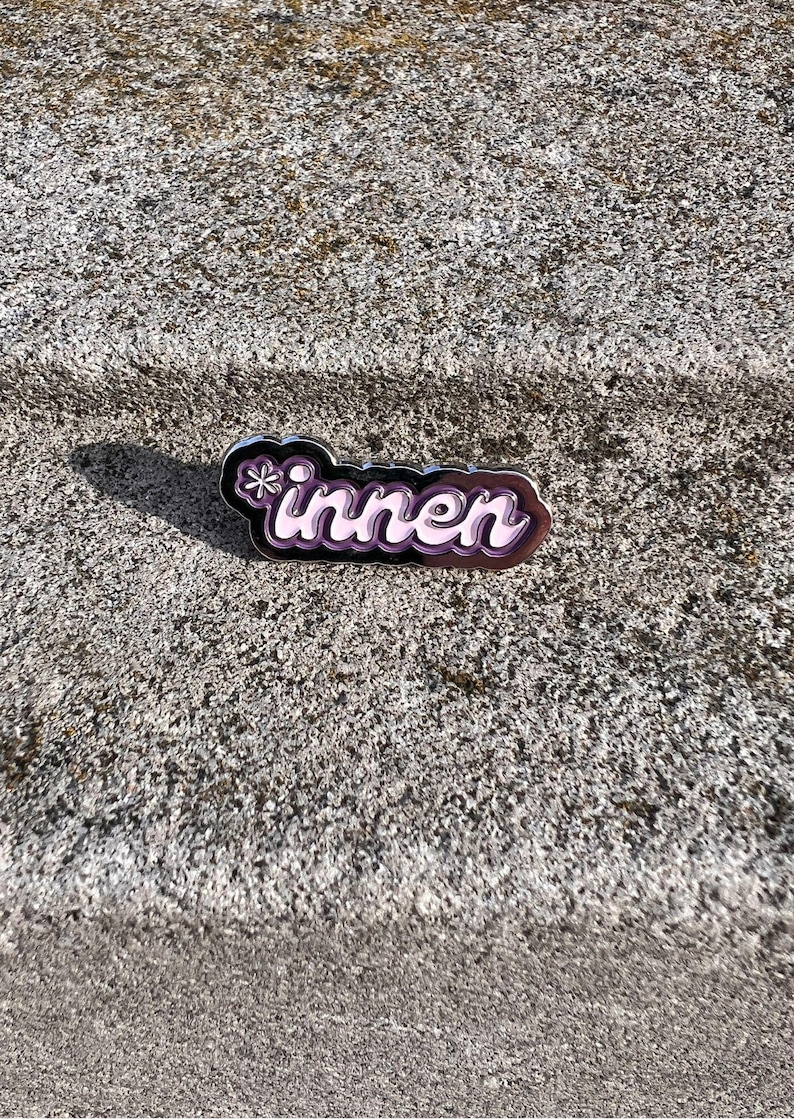 Op de afbeelding: Een paarse en witte emaille pin met het Duitse woord "innen" in een gestileerd lettertype. De pin heeft een zwarte omtrek en een klein bloemmotief aan de linkerkant. De pin is geplaatst op een getextureerd grijs oppervlak.