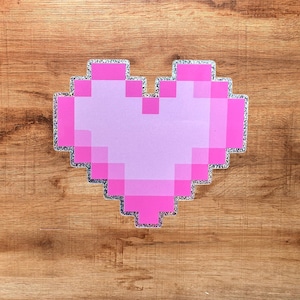 Könnte beinhalten: Ein pixeliger Herzaufkleber mit einem helllavendelfarbenen Zentrum und einem leuchtend pinken Rand. Die gesamte Herzform ist mit einer glitzernden silbernen Kante umrandet, die einen deutlichen Kontrast zur umgebenden Holzoberfläche bildet.