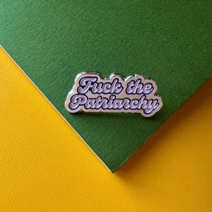 Könnte beinhalten: Ein silberfarbener Emaille-Anstecker mit dem Schriftzug "Fuck the Patriarchy" in Schreibschrift. Der Anstecker befindet sich auf einem grünen und gelben Hintergrund.