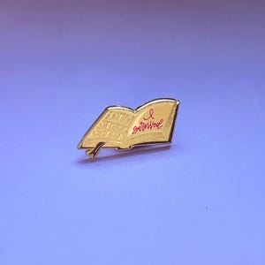 Könnte beinhalten: Eine goldfarbene Metall-Emaille-Anstecknadel in Form eines aufgeschlagenen Buches. Die linke Seite zeigt den geprägten Text "LONG STORY SHORT" und die rechte Seite den roten Schreibschrifttext "I survived" mit einer kleinen roten Schleife darüber.