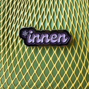 Op de afbeelding: Een paarse en witte emaille pin met het woord "innen" in een gestileerd lettertype, gezet tegen een zwarte rand. De pin heeft een klein bloemmotief. De achtergrond is een gele ruitvormige oppervlakte.