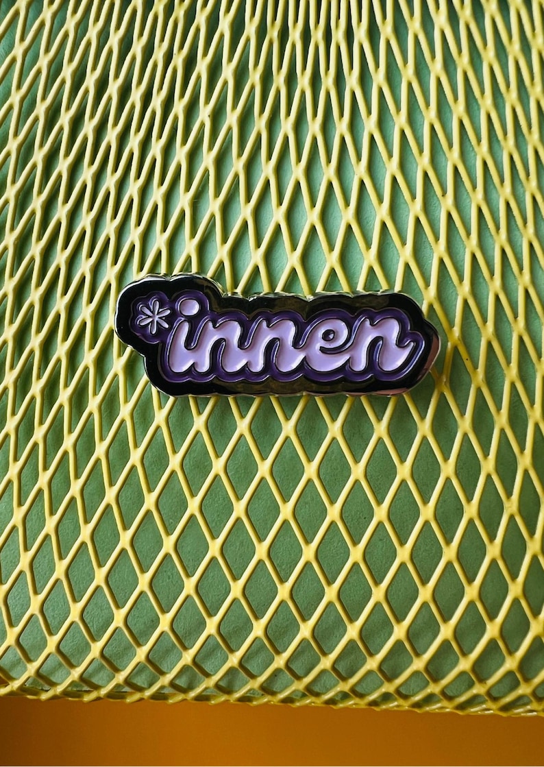 Op de afbeelding: Een paarse en witte emaille pin met het woord "innen" in een gestileerd lettertype, gezet tegen een zwarte rand. De pin heeft een klein bloemmotief. De achtergrond is een gele ruitvormige oppervlakte.
