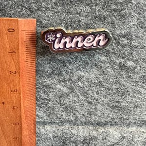 Op de afbeelding: Een zilverkleurige pin met het woord "innen" in een cursief lettertype, met een sneeuwvlokontwerp aan het begin. De letters zijn lichtroze met een donkerroze omlijning. De pin ligt op een grijze, getextureerde ondergrond.