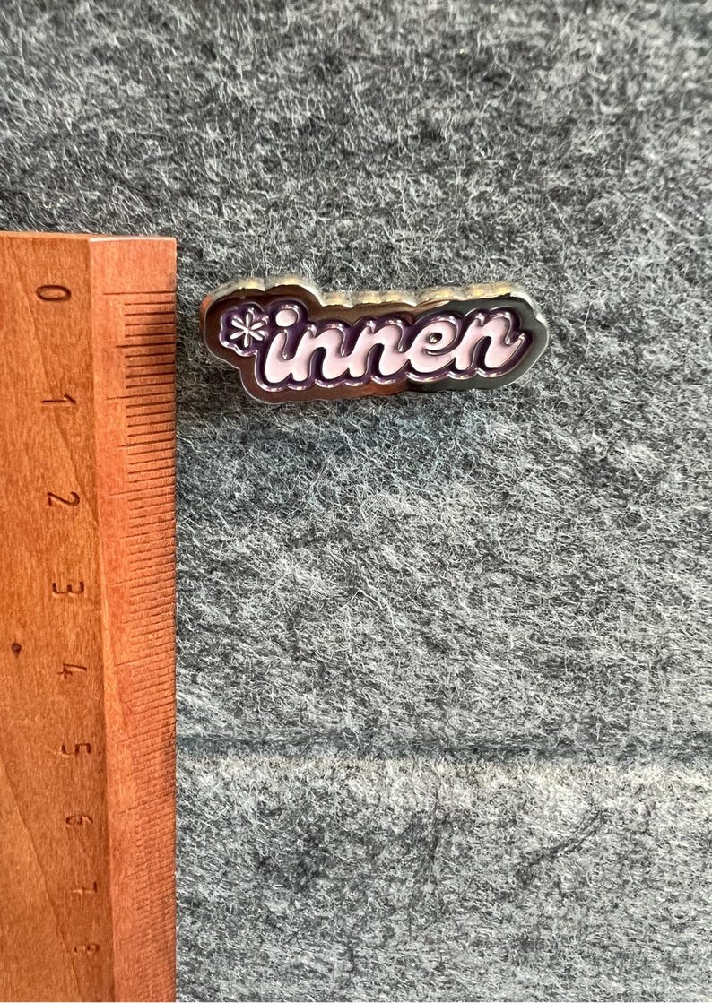Op de afbeelding: Een zilverkleurige pin met het woord "innen" in een cursief lettertype, met een sneeuwvlokontwerp aan het begin. De letters zijn lichtroze met een donkerroze omlijning. De pin ligt op een grijze, getextureerde ondergrond.