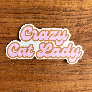 Puede incluir: Una pegatina con el texto "Crazy Cat Lady" en rosa con contorno marrón y fondo blanco. La pegatina está sobre una superficie de madera.