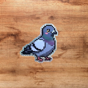 Puede incluir: Una pegatina holográfica con una ilustración en pixel art de una paloma. El ave aparece de perfil con plumaje en tonos grises, azules y morados, una mancha blanca en el ala y pequeñas patas rosas. La pegatina tiene un acabado brillante.
