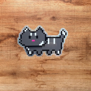 Op de afbeelding: Een glitterende sticker in pixel-artstijl van een donkergrijze kat met witte strepen op de rug en staart, een kleine roze mond en witte accenten op de oren en poten, weergegeven in een blokkerige, digitale esthetiek.