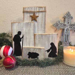 May include: A rustic Christmas nativity scene with wooden blocks, featuring black silhouette figures. A metal cross and a lit candle are also present. Red and white striped ornaments and a star-shaped decoration add festive touches.