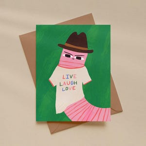 May include: Greeting card featuring a whimsical illustration of a pink worm-like creature wearing a brown hat and a white t-shirt with the words "LIVE LAUGH LOVE" in colorful letters. The card is set against a green background and comes with a brown envelope.