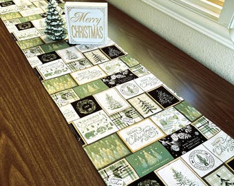 Pode incluir: Um caminho de mesa com tema natalino com um design de patchwork com quadrados verdes, pretos e brancos. Os quadrados exibem árvores de Natal, coroas e os textos "Merry Christmas" e "Season's Greetings". Uma pequena árvore de Natal e uma placa com "Merry Christmas" estão em cima.