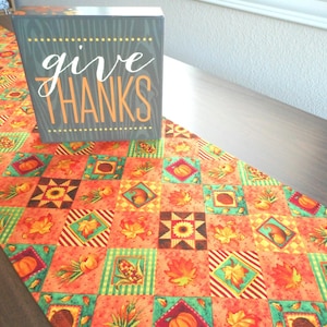 May include: A fall-themed table runner with a patchwork design featuring pumpkins, leaves, and acorns. The runner is orange, green, and brown. A wooden sign with the text "give thanks" is on top of the runner.
