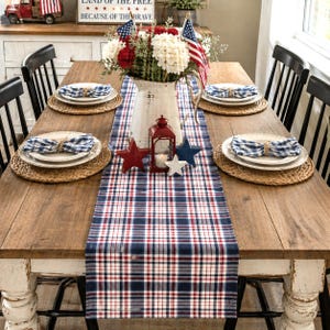 May include: A rustic wooden dining table set for a patriotic celebration. The table is adorned with a red, white, and blue plaid table runner, place settings, and decorative stars. A vase of flowers and American flags complete the festive look.
