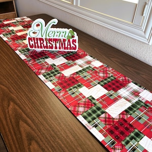 May include: A red and green plaid table runner with a "Merry Christmas" sign on top. The table runner is made of fabric and has a patchwork design.
