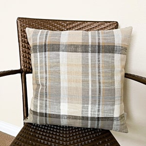 May include: A square throw pillow featuring a plaid pattern in shades of beige, tan, gray, and white. The fabric has a visible woven texture with intersecting horizontal and vertical lines forming a classic checkered design across the entire surface.