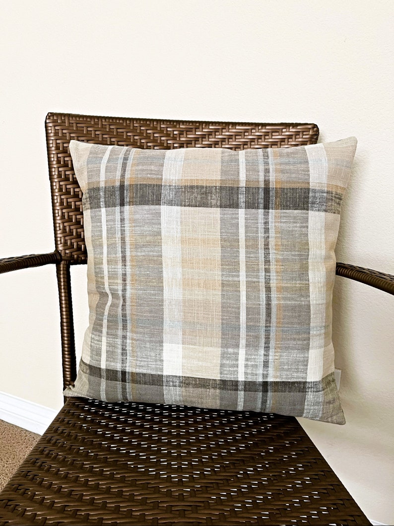 May include: A square throw pillow featuring a plaid pattern in shades of beige, tan, gray, and white. The fabric has a visible woven texture with intersecting horizontal and vertical lines forming a classic checkered design across the entire surface.