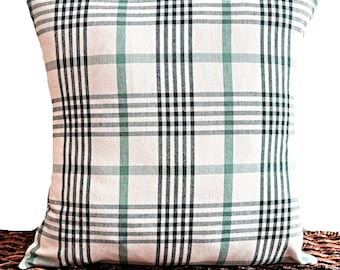 May include: Square throw pillow with a white background and a plaid pattern in shades of green and black. The pillow is resting on a woven brown surface. The pillow is a decorative home accent.