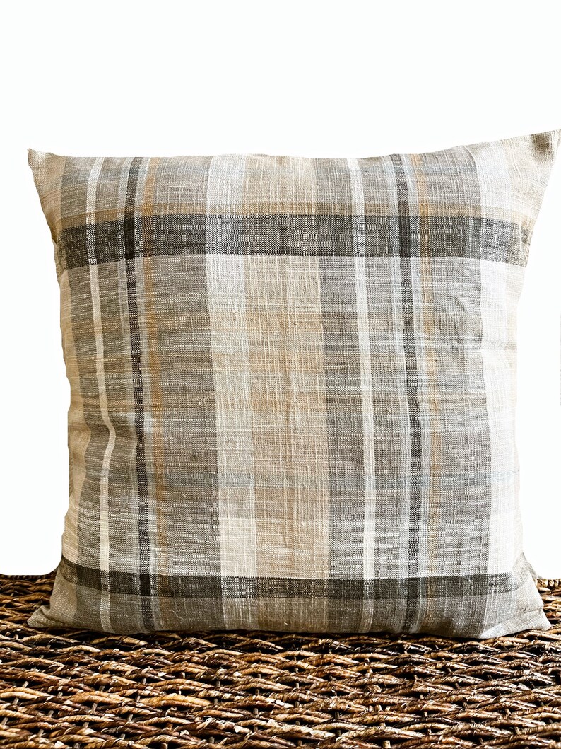 May include: A square throw pillow featuring a woven plaid pattern in neutral shades of beige, light gray, dark charcoal, and soft white. The fabric has a visible textured weave, and the design consists of intersecting vertical and horizontal stripes.