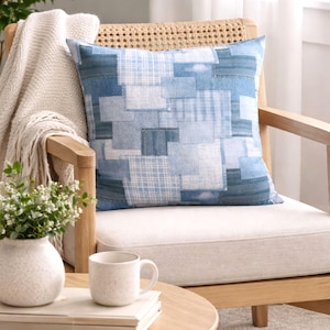 May include: A square denim patchwork pillow with various shades of blue and white sits on a light-colored chair. The pillow's design features a mix of denim squares and rectangles. A beige throw blanket is draped over the chair.
