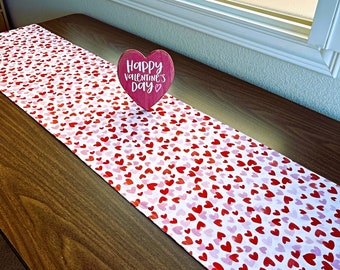 May include: A white table runner with a pink and red heart pattern. A pink wooden heart sign with the text "Happy Valentine's Day" is on top of the runner.